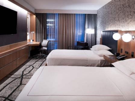 The image shows a modern hotel room with two beds, a TV, a desk, and stylish decor. The lighting is soft and inviting.