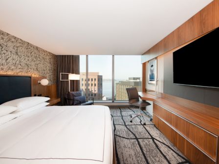 A modern hotel room with a large bed, wall-mounted TV, desk, and a window view of city buildings and water in the distance.
