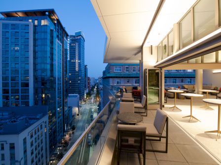 A modern rooftop terrace overlooking a city street, with chairs, tables, and buildings lit up during dusk.