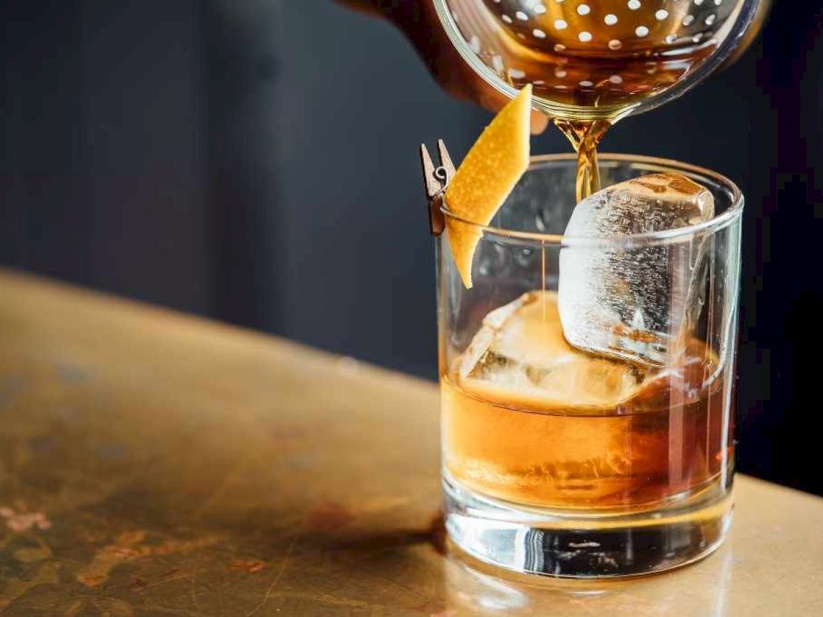A hand pours a drink into a glass with a large ice cube and a twist of citrus peel, sitting on a wooden surface.