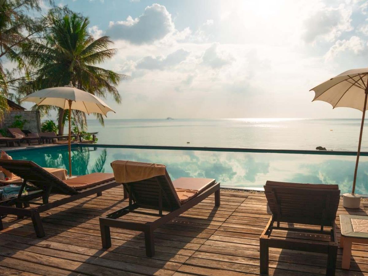 A serene beach scene with lounge chairs, umbrellas, and palm trees by a pool overlooking the ocean at sunset.