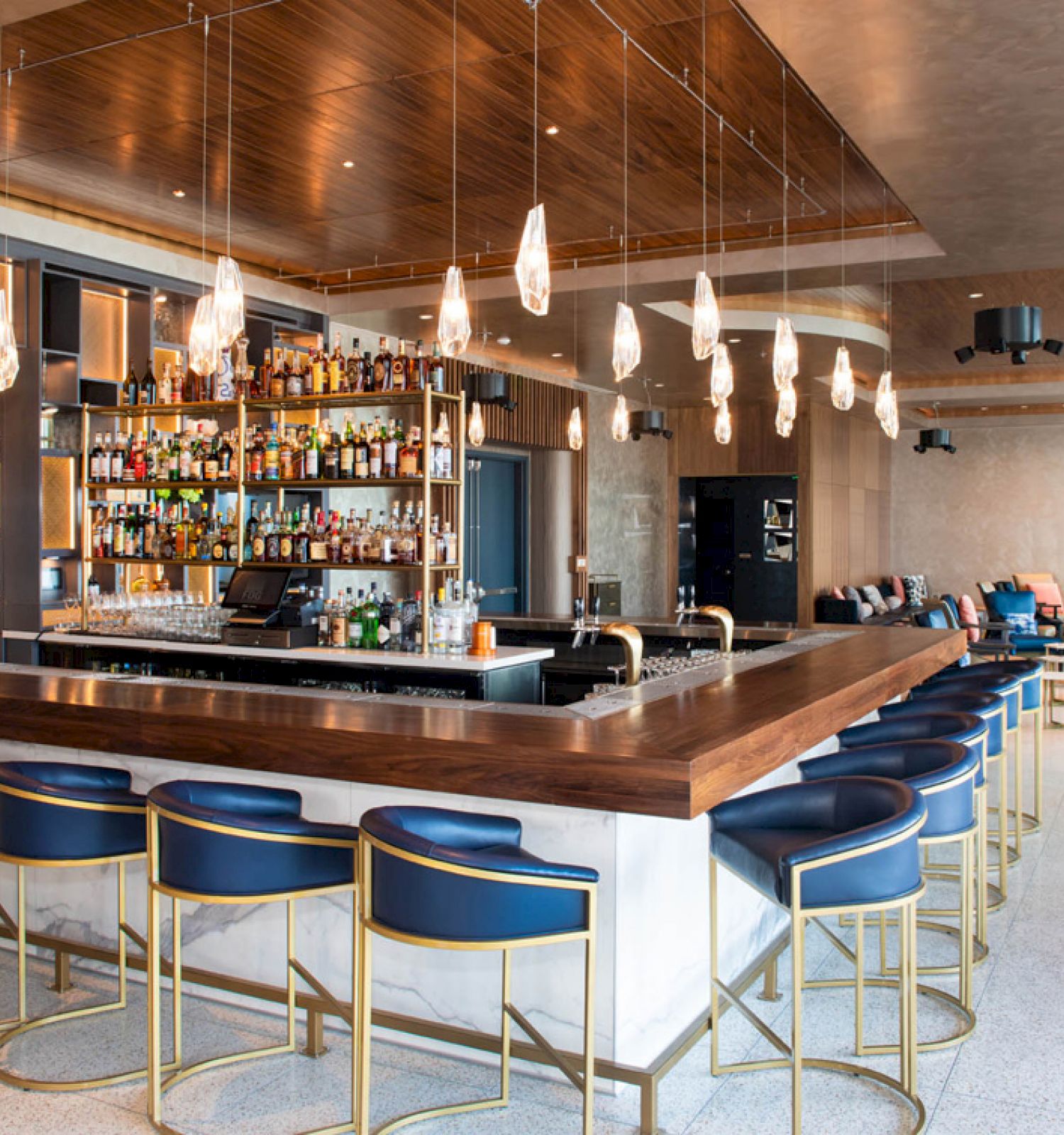The image shows a modern bar with stylish seating, hanging lights, and a well-stocked shelf of bottles. There's a cozy lounge area nearby.