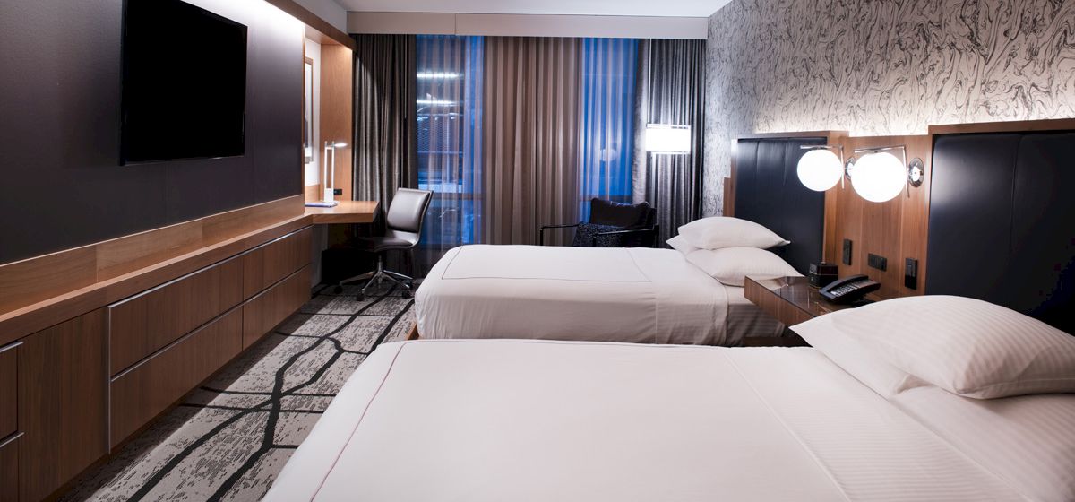 The image shows a modern hotel room with two beds, a TV, a desk, and a window with curtains, creating a cozy and stylish atmosphere.