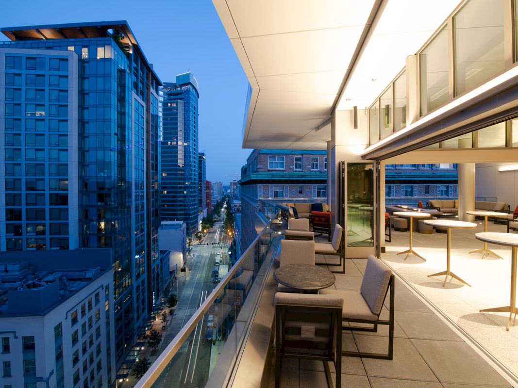 A modern cityscape with tall buildings is seen from a stylish outdoor balcony with furniture, overlooking a street at dusk.