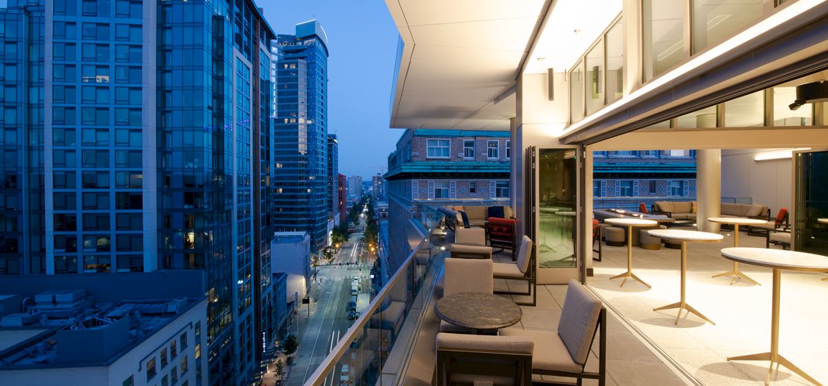 A modern cityscape with tall buildings is seen from a stylish outdoor balcony with furniture, overlooking a street at dusk.