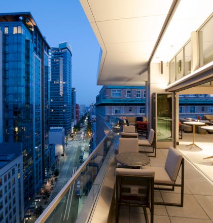 A modern cityscape at dusk features a furnished balcony overlooking tall buildings and a street below, with interior seating visible.