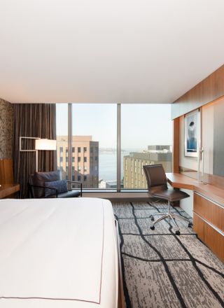 A modern hotel room with a large bed, a wall-mounted TV, a desk, and a window view of buildings and water in the background.