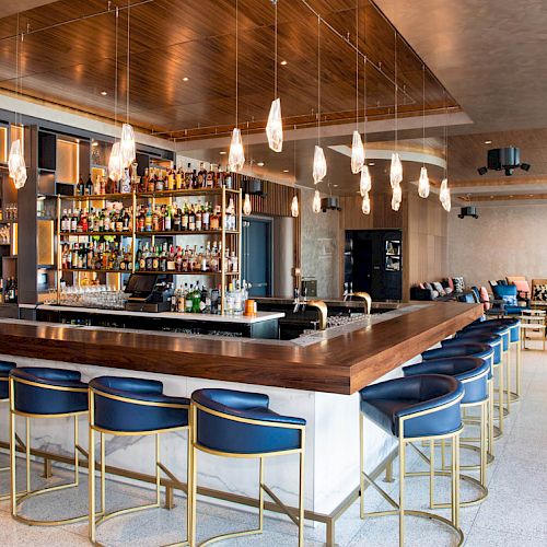 A modern bar with a wooden counter, blue stools, hanging lights, and a stocked liquor shelf in a stylish and spacious lounge area.