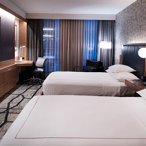 A modern hotel room with two beds, a desk, a TV, stylish lighting, and patterned wallpaper.