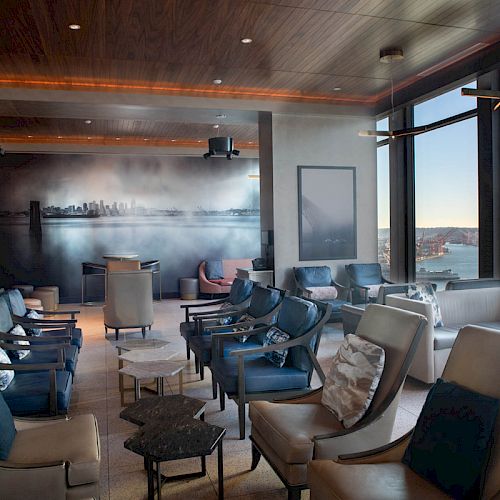 The image shows a modern lounge with plush seating, a city skyline mural, and large windows overlooking a waterfront view and Ferris wheel.