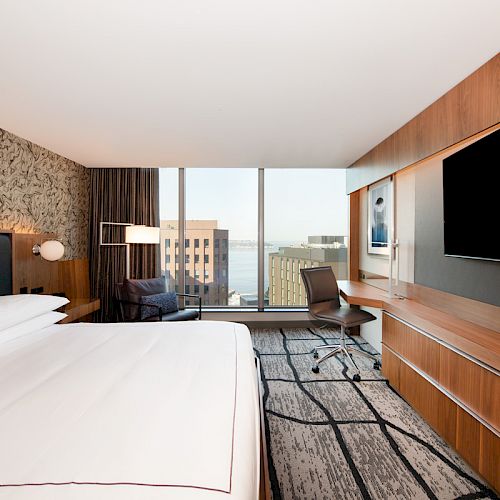 A modern hotel room with a large bed, wall-mounted TV, work desk, seating area, abstract wall art, and a window with a city view.