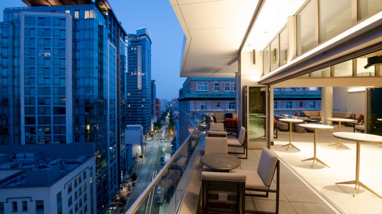 A modern rooftop balcony with a cityscape view, featuring seating, tables, and large glass windows in an urban setting.