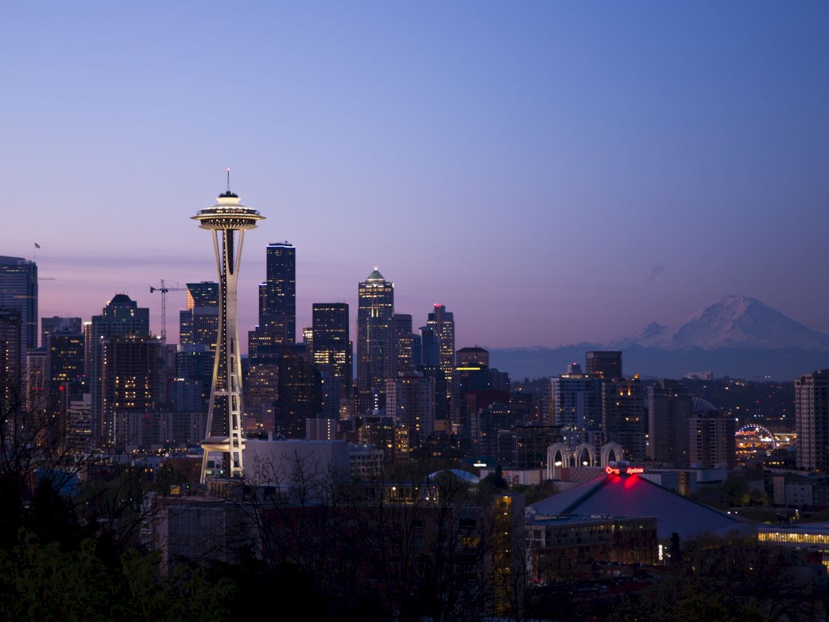Seattle skyline at dusk featuring the Space Needle and Mount Rainier in the background, with city lights illuminating the scene.