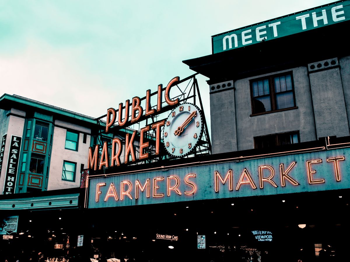 The image shows a sign for a "Public Market" and "Farmers Market" with a clock, set against some buildings with various signs.