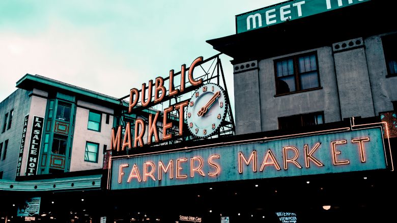 The image shows a sign for a "Public Market" and "Farmers Market" with a clock, set against some buildings with various signs.