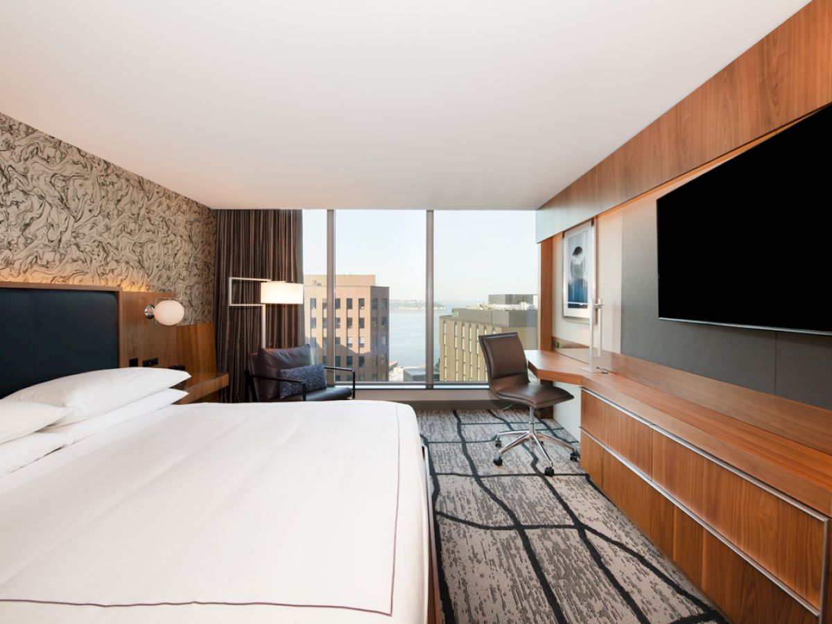 A modern hotel room with a large bed, TV, desk, and window offering a city view.