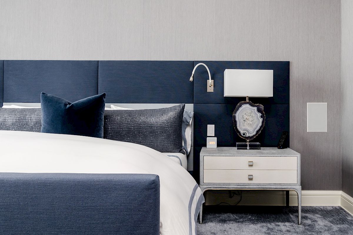 A modern bedroom with a blue headboard, side table, lamp, and gray carpet. Bedding is white with blue accents, creating a sleek look.