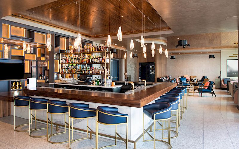 A modern bar with blue stools, hanging lights, and a well-stocked shelf. The space is stylish and inviting, with seating in the background.