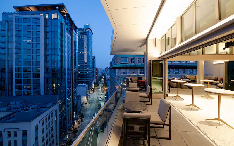 A modern rooftop terrace with seating, overlooking a cityscape of tall buildings at dusk, creating a vibrant urban atmosphere.