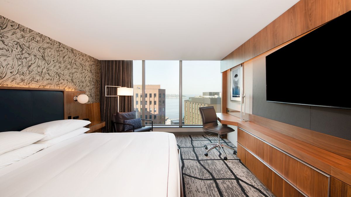A modern hotel room features a large bed, TV, desk, and chair, with a window view of buildings and water in the background.