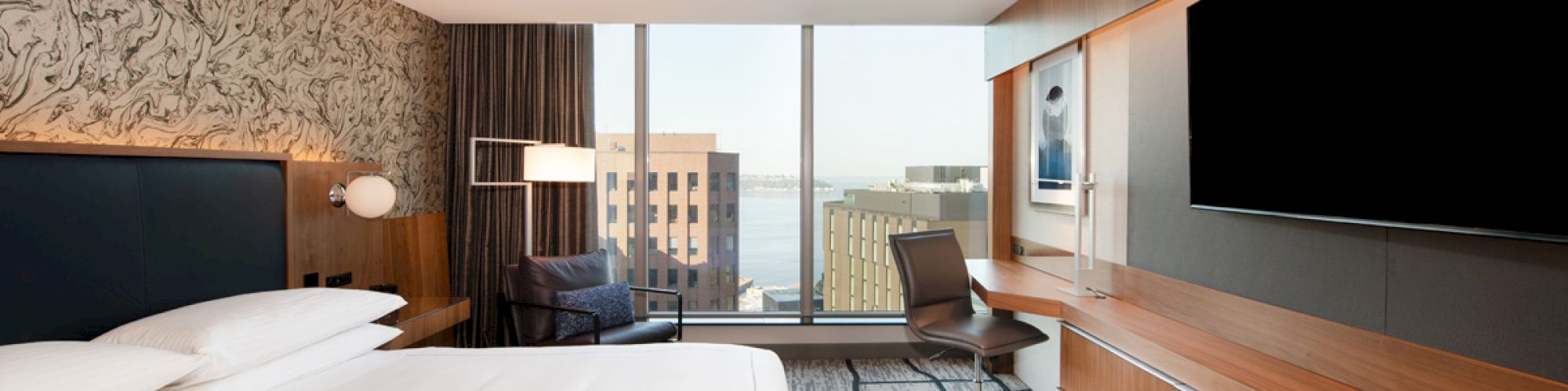 A modern hotel room features a large bed, TV, desk, and chair, with a window view of buildings and water in the background.